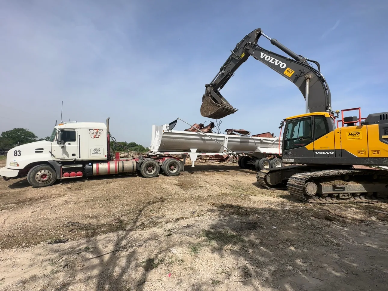 Excavator loading debris next to a lowboy haul truck on a cleared demolition site