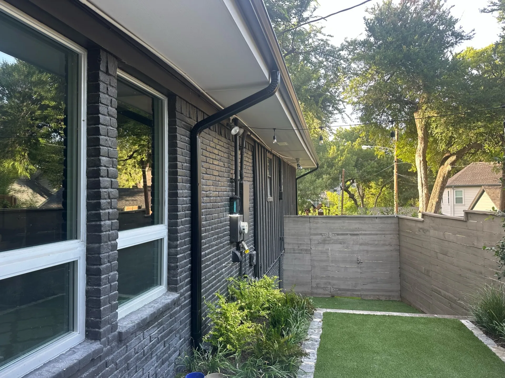 Gutter installation on a modern brick home in Lakewood, TX, showing sleek black downspouts, a landscaped backyard, and privacy fencing with turf lawn